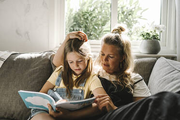 Daughter writing in book while sitting by mother on sofa at home - MASF21594