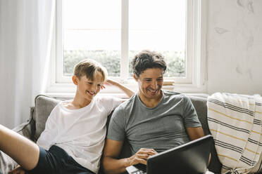 Happy father using laptop while sitting by son in living room - MASF21603