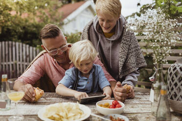 Younger brother using mobile phone sitting by father and sibling at table in dinner party - MASF21609