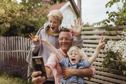 Fröhlicher Vater nimmt Selfie mit Söhnen auf Smartphone im Vorgarten am Abend - MASF21611