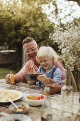 Son using mobile phone sitting with father in back yard - MASF21614