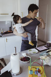 Father eating food while baby son looking at him in kitchen at home - MASF21679