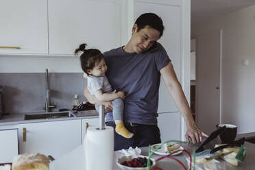 Father talking on smart phone while using digital tablet while carrying baby son in kitchen - MASF21680