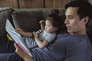 Vater mit männlichem Kleinkind, der auf dem Sofa im Wohnzimmer liegt und ein Buch liest - MASF21695