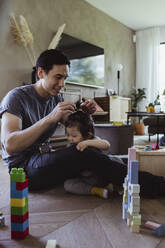 Smiling father tying hair of male toddler's at home - MASF21700