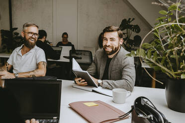Lächelnder Geschäftsmann, der einen Terminkalender hält, während er mit einem männlichen Kollegen in einem Coworking-Büro sitzt - MASF21709
