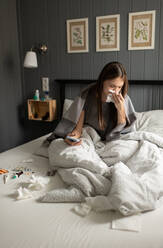 Sick female with smartphone sneezing on bed - CAVF92130