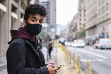 Junger Mann mit Schutzmaske, der ein Mobiltelefon benutzt, während er in der Stadt steht - AGOF00001