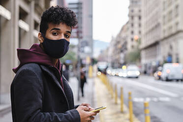 Young man wearing protective face mask using mobile phone while standing in city - AGOF00001