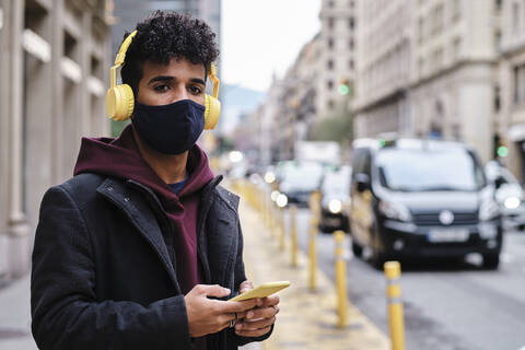 Mann mit Jacke und Gesichtsschutzmaske, der ein Mobiltelefon benutzt, während er auf der Straße in der Stadt steht, lizenzfreies Stockfoto