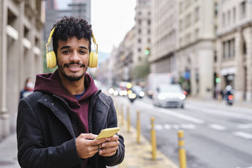 Lächelnder Mann mit Kopfhörern, der ein Smartphone benutzt, während er auf der Straße in der Stadt steht - AGOF00004