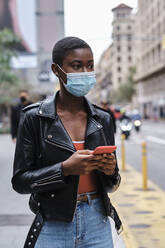 Young woman wearing protective face mask using mobile phone while standing in city - AGOF00005