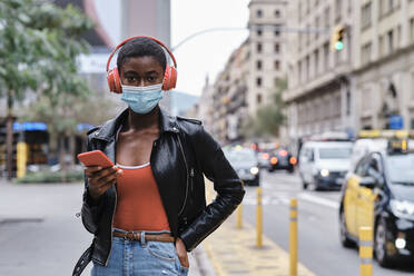 Woman wearing protective face mask and headphones using mobile phone while standing  - AGOF00006