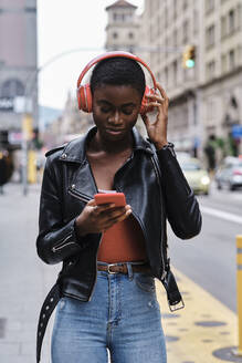 Woman listening music through headphones while using smart phone standing in city - AGOF00010