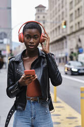 Young woman with mobile phone listening music through headphones while standing in city - AGOF00011