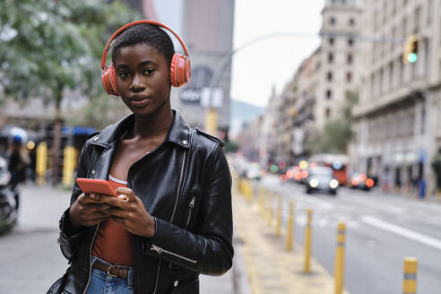 Woman wearing headphones and jacket using smart phone while standing in city - AGOF00012