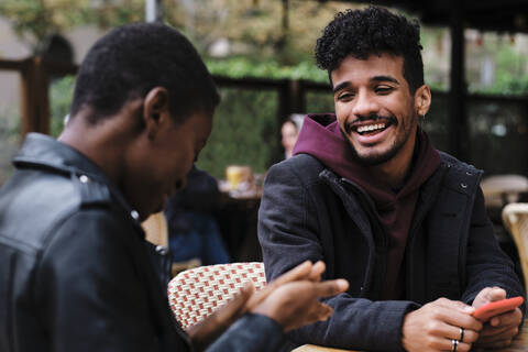 Lächelnder Mann, der einen Freund ansieht, während er in einem Straßencafé sitzt, lizenzfreies Stockfoto