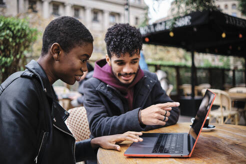 Man pointing toward laptop while sitting by friend at sidewalk cafe - AGOF00016