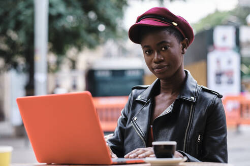 Young woman wearing jacket and cap using laptop while sitting at sidewalk cafe in city - AGOF00019