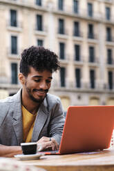 Fashionable man smiling while working on laptop sitting at side walk cafe - AGOF00022