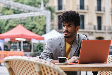 Junger, modischer Mann mit Laptop, der wegschaut, während er in einem Straßencafé sitzt, lizenzfreies Stockfoto