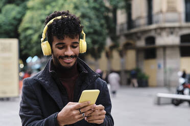 Lächelnder Mann mit Kopfhörern, der ein Mobiltelefon benutzt, während er in der Stadt steht - AGOF00028
