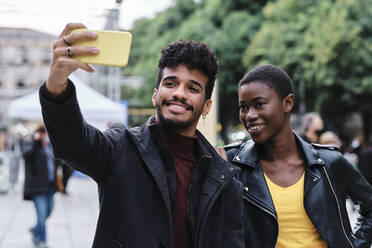 Smiling man taking selfie with friend through mobile phone while standing in city - AGOF00029