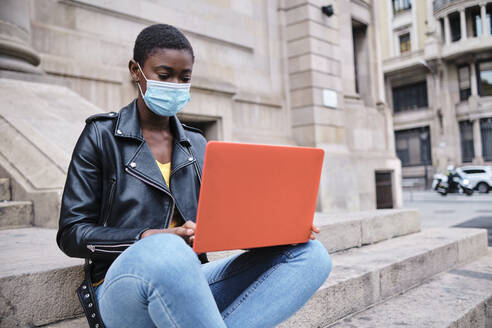 Young woman wearing protective face mask using laptop while sitting on steps in city - AGOF00032