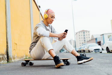 Junge Frau, die auf einem Skateboard sitzt, Kopfhörer trägt und ein Mobiltelefon benutzt - ISF24348