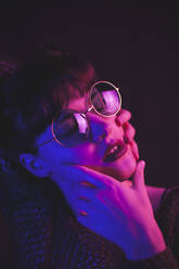 Ruhige Frau mit stilvoller Brille, die ihr Gesicht berührt, während sie mit geschlossenen Augen auf dunklem Hintergrund in rosa und blauer Neonbeleuchtung steht - ADSF20652