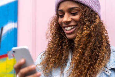 Fröhlicher afroamerikanischer weiblicher Teenager in modischem Outfit und Hut, der lächelnd eine Nachricht liest, während er auf der Straße in der Nähe einer rosa Tür steht - ADSF20657