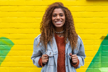 Optimistic young black female millennial with curly hair in stylish clothes smiling while looking at camera against yellow wall - ADSF20658