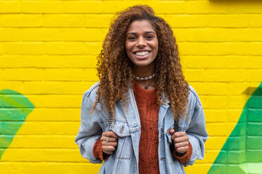 Optimistische junge schwarze Millennials mit lockigem Haar in modischer Kleidung lächelnd in die Kamera blickend vor einer gelben Wand - ADSF20658
