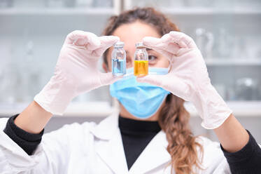 Junge Wissenschaftlerin mit Gesichtsmaske, die zwei blaue und orangefarbene Fläschchen mit Coronavirus-Impfstoff in der Hand hält. Covid-19-Impfstoffentwicklung. - CAVF92258