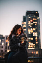 Teenage girl using smart phone sitting on railing in city at night - CAVF92281