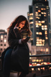 Teenager-Mädchen mit Smartphone sitzt auf Geländer in der Stadt bei Nacht - CAVF92282