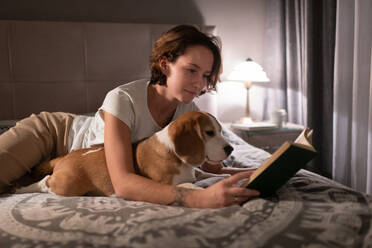 Junge Frau mit Beagle, die auf dem Bett ein Buch liest - CAVF92302