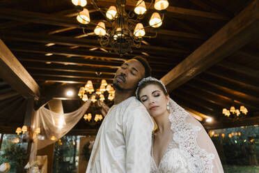 Bride and groom wearing wedding dress leaning on each other while standing at banquet - EGAF01675