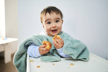 Niedliches Baby mit Handtuch isst Croissant, während es zu Hause am Tisch sitzt - KIJF03546