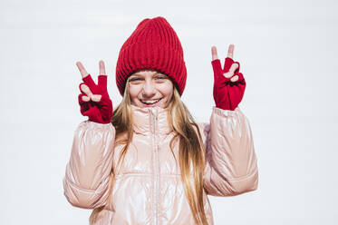 Smiling girl in warm clothing showing Peace Sign against wall - JCMF01863