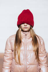 Girl in wall clothing covering eyes through knit hat against white wall - JCMF01871