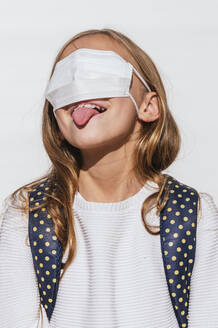 Playful girl sticking out tongue while covering eyes with protective face mask during COVID-19 - JCMF01874