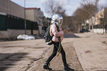 Mädchen mit Weltraumhelm benutzt Mobiltelefon beim Gehen auf der Straße - JCMF01878
