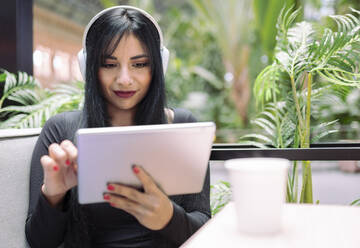 Schöne junge Frau hört Musik, während sie ein digitales Tablet im Café benutzt - JCCMF01189