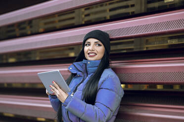 Frau in warmer Kleidung mit digitalem Tablet, die an einem Geländer steht und wegschaut - JCCMF01193