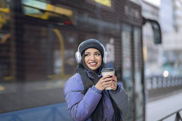 Smiling young woman with headphones holding coffee cup in city - JCCMF01197