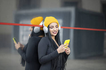 Schöne Frau mit Kopfhörern über einer Strickmütze, die ihr Smartphone gegen ein Glas hält - JCCMF01201