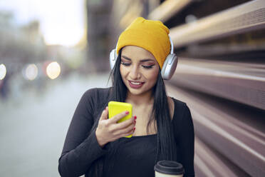 Junge schöne Frau hört Musik über Kopfhörer, während sie ein Smartphone auf der Straße benutzt - JCCMF01202