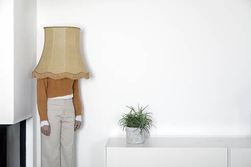 Ältere Frau mit Lampenschirm auf dem Kopf steht gegen eine weiße Wand - PSTF00832