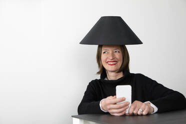 Glückliche Frau mit schwarzem Lampenschirm und Smartphone schaut weg - PSTF00839